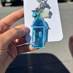Puede incluir: Un llavero de resina brillante azul y verde con forma de edificio. El llavero tiene un llavero y dijes plateados, incluyendo una mariposa, un corazón y una cuenta. El llavero está sujeto a una tarjeta blanca.