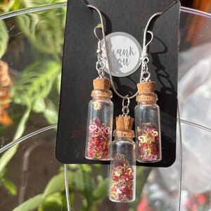Puede incluir: Un conjunto de collar y pendientes con botellas de vidrio en miniatura llenas de confeti rojo y dorado en forma de corazón. Las botellas tienen tapones de corcho y están suspendidas de cadenas plateadas. El fondo incluye una tarjeta negra con la frase "thank you".