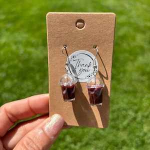 Puede incluir: Pendientes de café helado en miniatura con vasos de plástico transparente y líquido marrón oscuro. Los pendientes están sujetos a una tarjeta marrón con una pegatina de "Thank you".
