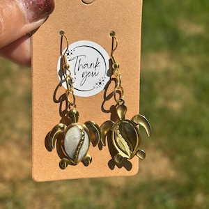 Puede incluir: Un par de pendientes de tortuga en tono dorado sujetos a una tarjeta de presentación rectangular marrón. Los dijes de tortuga tienen un acabado dorado metálico con una incrustación central, una blanca y otra marrón oscuro. La tarjeta incluye una pegatina circular con el texto Thank you.