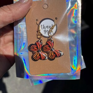 Puede incluir: Pendientes de mariposa con alas naranjas, negras y blancas, presentados en una etiqueta marrón con las palabras "thank you". Los pendientes cuelgan de ganchos dorados y están en una bolsa holográfica.