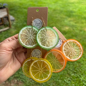 Puede incluir: Pendientes hechos a mano con forma de rodajas de cítricos. Los pendientes presentan diseños de lima verde, limón amarillo y naranja. Cada pendiente tiene un gancho dorado y una pequeña etiqueta que dice "Thank You".