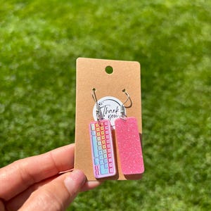 Puede incluir: Un par de pendientes colgantes en una tarjeta marrón. Un pendiente tiene un diseño de teclado colorido, el otro es un rectángulo rosa con purpurina. La tarjeta tiene un pequeño círculo con la frase "Thank you".