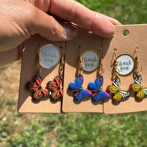 Puede incluir: Tres pares de pendientes colgantes con forma de mariposa en tarjetas de presentación de cartón marrón. Cada par tiene ganchos dorados y una pegatina circular que dice thank you. Las mariposas son naranja y negro, azul y morado, y amarillo y negro.