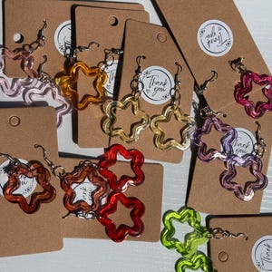 Puede incluir: Una colección de pendientes colgantes con forma de estrella en varios colores translúcidos, incluyendo rosa, amarillo, naranja, rojo, morado y verde lima. Cada par está sujeto a una tarjeta rectangular marrón con una pegatina que dice Thank you.