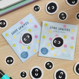 May include: A set of iron-on patches featuring black and white soot sprites. The patches are on a blue background with colourful stars and swirls. The text "SOOT SPRITES IRON-ON PATCH" is printed on the patches.