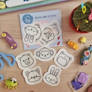 Può includere: Una collezione di materiali per l'artigianato e oggetti decorativi, tra cui toppe di animali "Stick and Stitch", figurine in ceramica colorate, filo da ricamo e una tastiera blu. Le toppe presentano simpatici volti di animali.