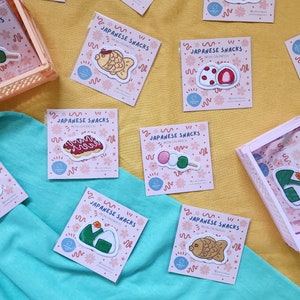 Peut inclure: Une collection de patchs thermocollants représentant différents en-cas japonais, dont des taiyaki en forme de poisson, des mochi, des dango et des onigiri. Les patchs sont sur un fond rose avec du texte blanc qui dit "Japanese Snacks Iron-on Patch".