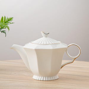 May include: White porcelain teapot with a gold trim and a lid with a small bird finial. The teapot has a octagonal body and a spout on the left side.