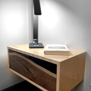 Può includere: Un comodino moderno sospeso con finitura in legno chiaro e frontale del cassetto in legno scuro. Una lampada da scrivania nera e un libro sono appoggiati sopra. Il comodino è montato su una parete bianca.