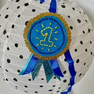May include: A white paper lantern with black polka dots features a blue felt award badge with a gold glitter scalloped border. The badge displays a yellow embroidered number one surrounded by stitched rays and hangs from blue satin ribbons.