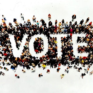 May include: An overhead shot shows a large group of people arranged to spell out the word "VOTE" on a white background. The individuals are diverse in appearance and clothing, creating a visual representation of community and civic engagement.