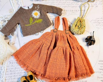 May include: A doll-sized ensemble featuring a brown long-sleeved sweater with a daisy design, an orange dress with suspenders, a small yellow purse, and tiny yellow shoes. The dress has a fringed hem.