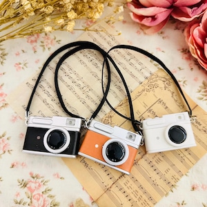 May include: Three miniature cameras with black straps. The cameras are black, orange, and white. The cameras are on a sheet of music.