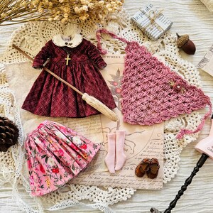 May include: A doll outfit set with a burgundy dress, a pink floral skirt, pink socks, and a pink crocheted shawl. The dress has a white collar and a gold cross necklace. The set is laid out on a white lace tablecloth.