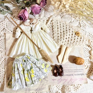 Könnte beinhalten: Ein Miniatur-Puppen-Outfit mit einem cremefarbenen Kleid mit Kreuzanhänger, einem floralen Rock, braunen Schuhen und einem Besen. Das Ensemble beinhaltet Socken und ist mit Trockenblumen und Vintage-Papier arrangiert.