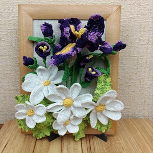 May include: A framed crocheted wall art piece featuring a purple iris flower with yellow accents and white daisies with yellow centers. The flowers are surrounded by green crocheted leaves.