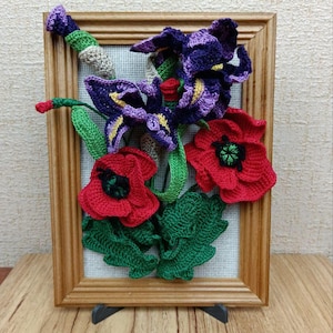 May include: A framed crocheted artwork featuring a bouquet of red poppies and purple irises. The flowers are crafted with yarn and displayed on a beige canvas.