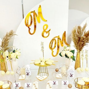 Puede incluir: Una mesa de postres de boda con detalles dorados. La mesa presenta un soporte para pastel dorado con cake pops blancos, candelabros geométricos dorados y flores blancas. El fondo es una pared blanca con letras dorées que deletrean "Mr & Mrs".