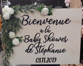 May include: A wooden sign with white flowers and greenery around the edges. The sign says "Bienvenue à la Baby Shower de Stéphanie 03.11.2019".