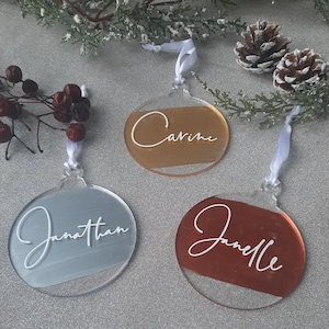 Może przedstawiać: Trzy okrągłe, przezroczyste ozdoby akrylowe z tłem w kolorze złotym, srebrnym i miedzianym. Każda ozdoba ma białą wstążkę i imię napisane białym pismem kursywą: "Carine", "Jonathan" i "Janelle".