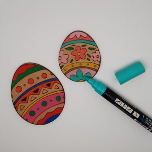 Puede incluir: Dos huevos de Pascua de madera, uno con un diseño floral rosa, verde y naranja y el otro con un diseño geométrico rosa, verde, azul y naranja. Un marcador turquesa está sobre la superficie blanca junto a los huevos.