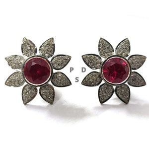 Puede incluir: Un par de pendientes de plata en forma de flor con una piedra preciosa roja en el centro de cada flor. Las flores están rodeadas de pequeños diamantes blancos.