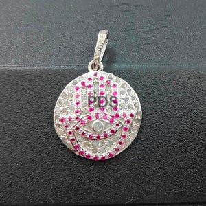 Puede incluir: Colgante plateado con un diseño circular que presenta un motivo estilizado de ojo y mano. El colgante está incrustado con pequeñas piedras transparentes y gemas rosas vibrantes. Las letras "PDS" son visibles.