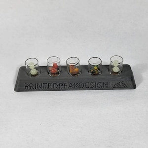 May include: Five small glass vials with cork stoppers, each containing a different colored bead or small object, are displayed on a black 3D printed stand with the text "PRINTEDPEAKDESIGN" printed on the front.