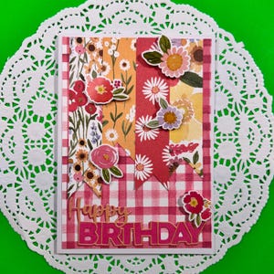 Può includere: Un biglietto d'auguri rettangolare con uno sfondo a quadretti vichy rosa e bianco e strisce verticali di carta con motivi floreali. Il biglietto è decorato con ritagli di fiori di carta sovrapposti e il testo Happy BIRTHDAY in rosa.