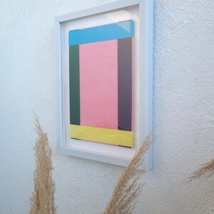 May include: A framed abstract art print with a pink background and colourful geometric shapes in blue, green, yellow, and purple.