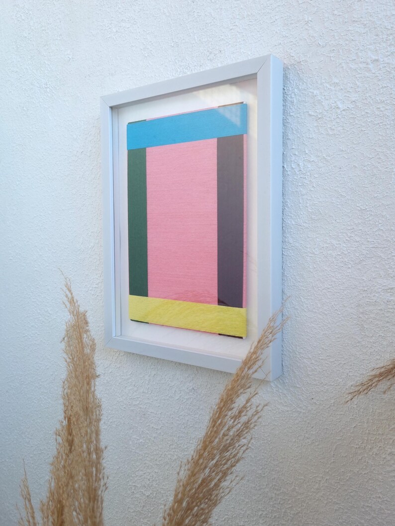 May include: A framed abstract art print with a pink background and colourful geometric shapes in blue, green, yellow, and purple.