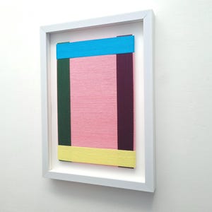 May include: A framed abstract art piece with a pink background and colourful stripes of blue, yellow, green, and purple.
