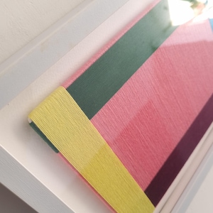 May include: A framed artwork with a geometric design made of colourful yarn. The artwork features a yellow, green, pink, and purple colour scheme.
