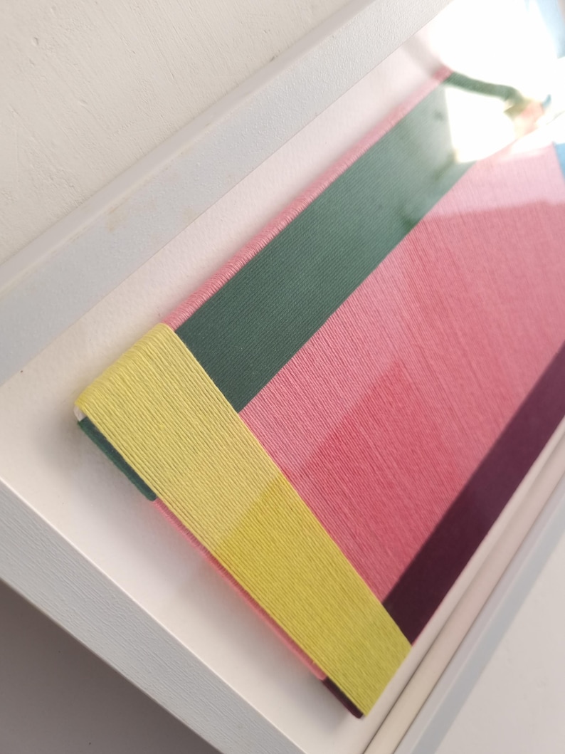 May include: A framed artwork with a geometric design made of colourful yarn. The artwork features a yellow, green, pink, and purple colour scheme.