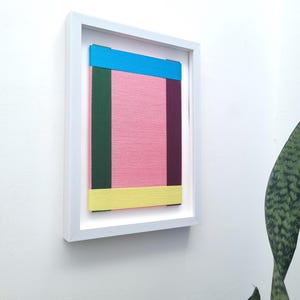 May include: A framed abstract art piece with a pink background and colourful stripes in blue, green, yellow, and purple.