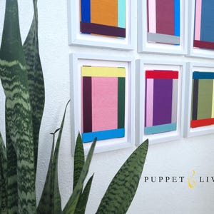 May include: A wall gallery of nine framed abstract paintings with geometric shapes and colourful stripes in shades of pink, blue, green, yellow, red, orange, and purple. The frames are white and the wall is white.