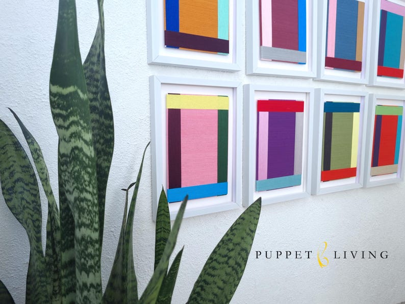 May include: A wall gallery of nine framed abstract paintings with geometric shapes and colourful stripes in shades of pink, blue, green, yellow, red, orange, and purple. The frames are white and the wall is white.