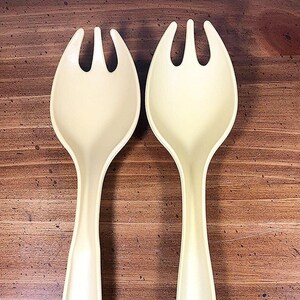 May include: A set of two off-white plastic salad servers with three prongs on each. The servers are on a wooden surface.