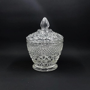 May include: A clear, lidded glass jar with a diamond pattern. The jar has a round base and a decorative finial on the lid. The glass is transparent, allowing light to pass through. The jar is likely used for storing small items or as a decorative piece.