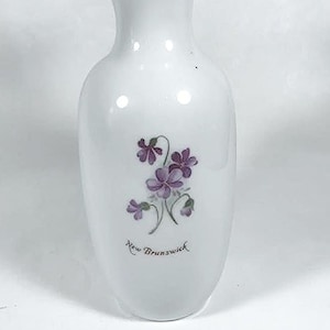 May include: White ceramic vase with a floral design of purple flowers and green leaves. The vase has the text "New Brunswick" written on the front.