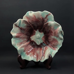 May include: A ceramic flower-shaped dish with a teal and burgundy glaze. The dish is on a wooden stand.