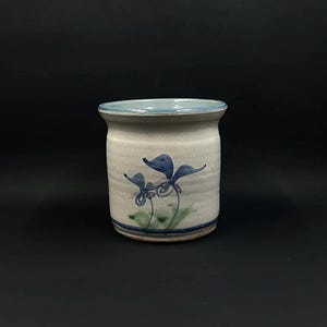 May include: A white ceramic utensil holder with a blue and purple floral design. The holder has a light blue rim and a white glaze.