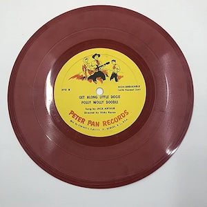 May include: A red vinyl record with a yellow label. The label features a cartoon image of three cowboys on horseback. The text on the label reads "Get Along Little Dogie Polly Wolly Doodle" and "Peter Pan Records".