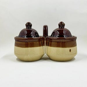 May include: A two-section ceramic condiment server with a handle. The server has a dark brown glaze on the lids and upper portion, with a tan band and base. The lids have a small knob on top.