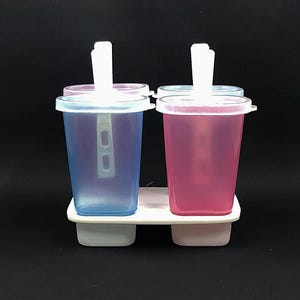 May include: A white plastic popsicle mold with four compartments. Two compartments are filled with blue liquid and two compartments are filled with pink liquid. Each compartment has a white plastic stick.