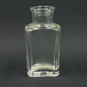 May include: A clear, rectangular glass bottle with a square base and a rounded neck. The bottle has a slightly textured surface and is empty. The bottle is likely vintage or antique.