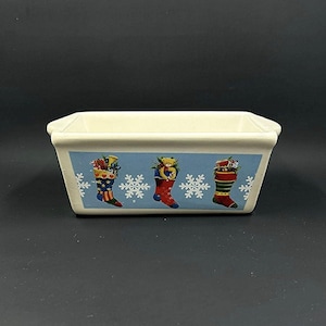 May include: Rectangular ceramic dish with a light cream color. The front features a blue band with illustrations of Christmas stockings filled with gifts and snowflakes. The dish is likely for serving food or as a decorative piece.