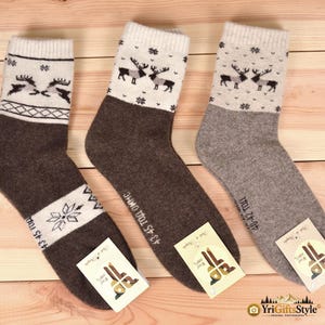 May include: Three pairs of wool socks featuring a cream-colored cuff with a dark brown or grey reindeer and snowflake pattern. The socks are solid dark brown or grey with additional white bands featuring snowflake designs and size text 43-45 or 40-42.