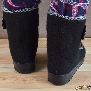 May include: A pair of black felt boots featuring a white zigzag stitch along the top edge, a black rubber sole, and a side closure strap. The interior is lined with white fleece, and the boots are worn with dark blue pants with a red paisley pattern.